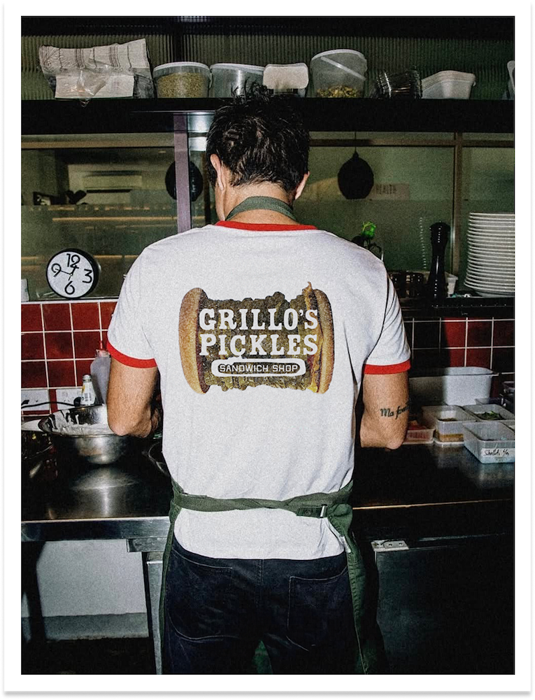 A person working in a sandwich shop, wearing a white t-shirt with red trim, with a logo that says "Grillo's Pickles Sandwich Shop" and an illustration of a pickle in the shape of a hot dog. The person is standing behind a kitchen counter with various containers and utensils, with a red-tiled wall and a clock showing the time as 9:23 in the background.