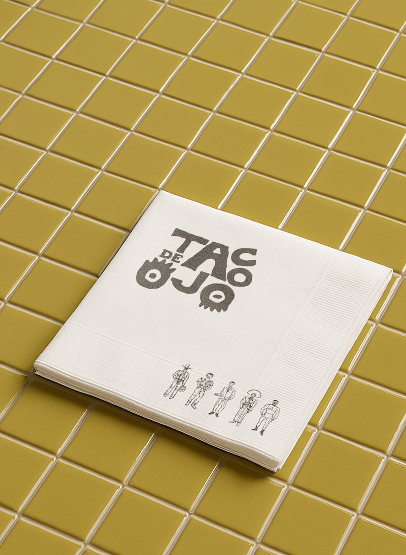 A white napkin with the logo "TAC DE JO" in black and a small illustration of six people in black line drawing at the bottom, resting on yellow tile flooring.