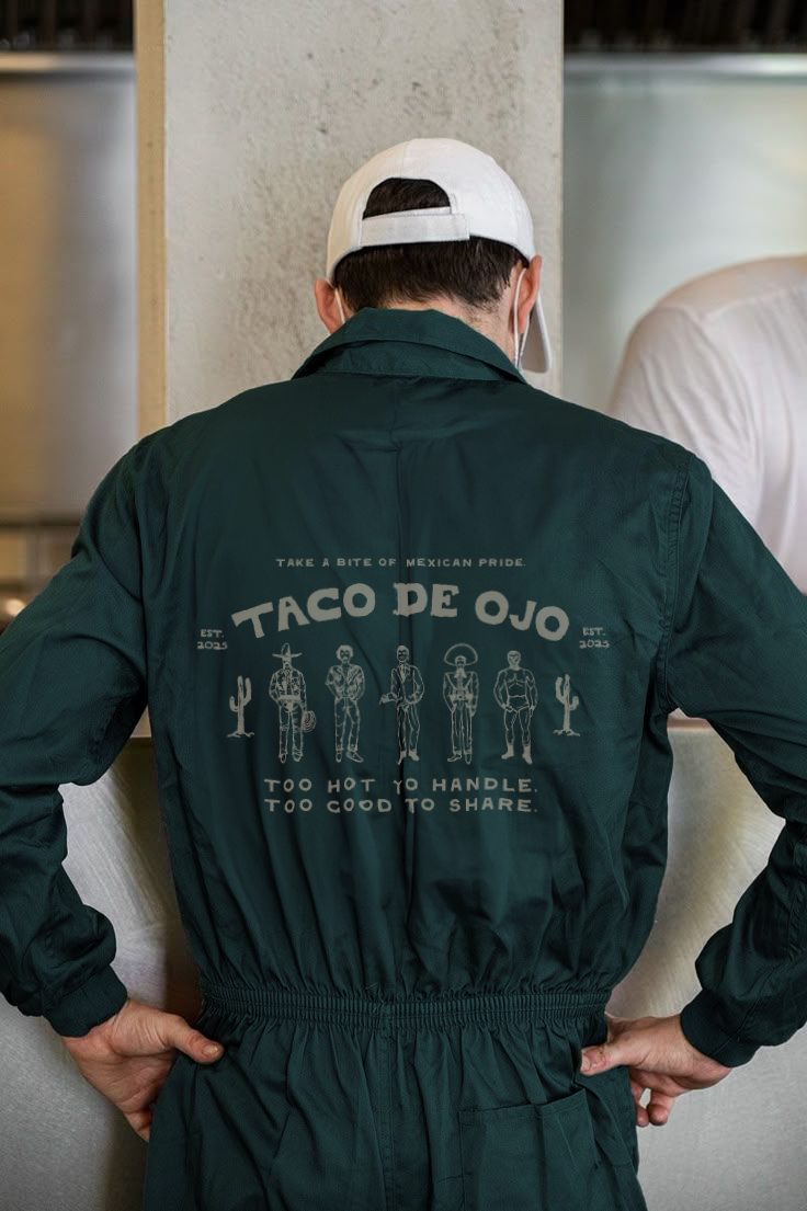 Back of a person wearing a dark green jacket with white text and illustrations, and a white baseball cap, sitting indoors.