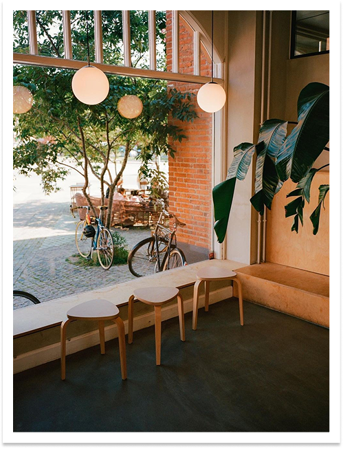 Interior of a café with three wooden stools at a large window, green plants, and bicycles outside.