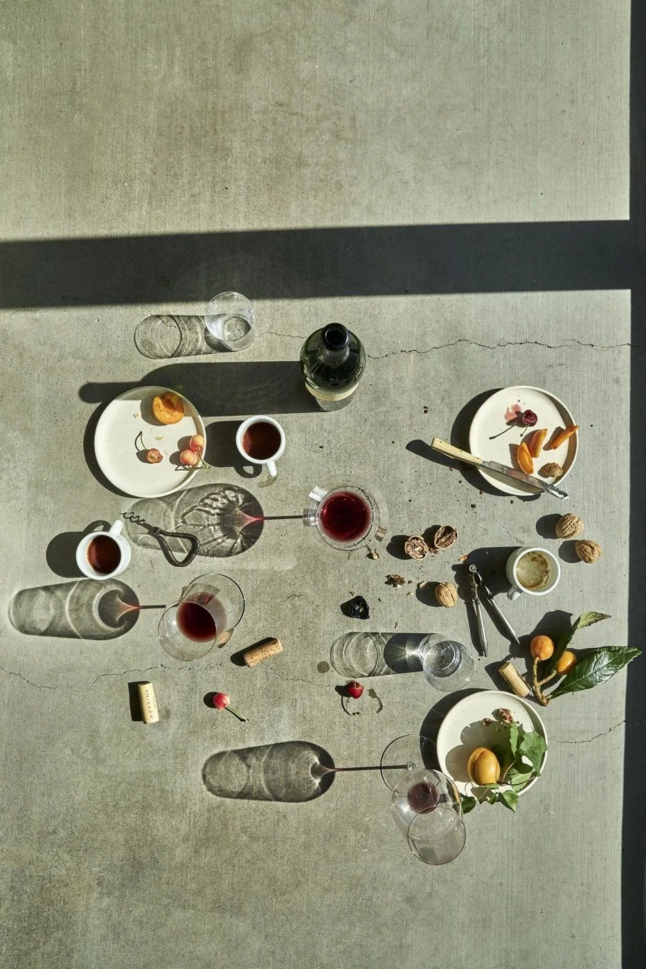 Top-down view of a table with wine glasses, empty plates with food scraps, a bottle of wine, a cork, a corkscrew, walnuts, cherries, and a small branch with leaves and fruit, illuminated by sunlight creating shadows.