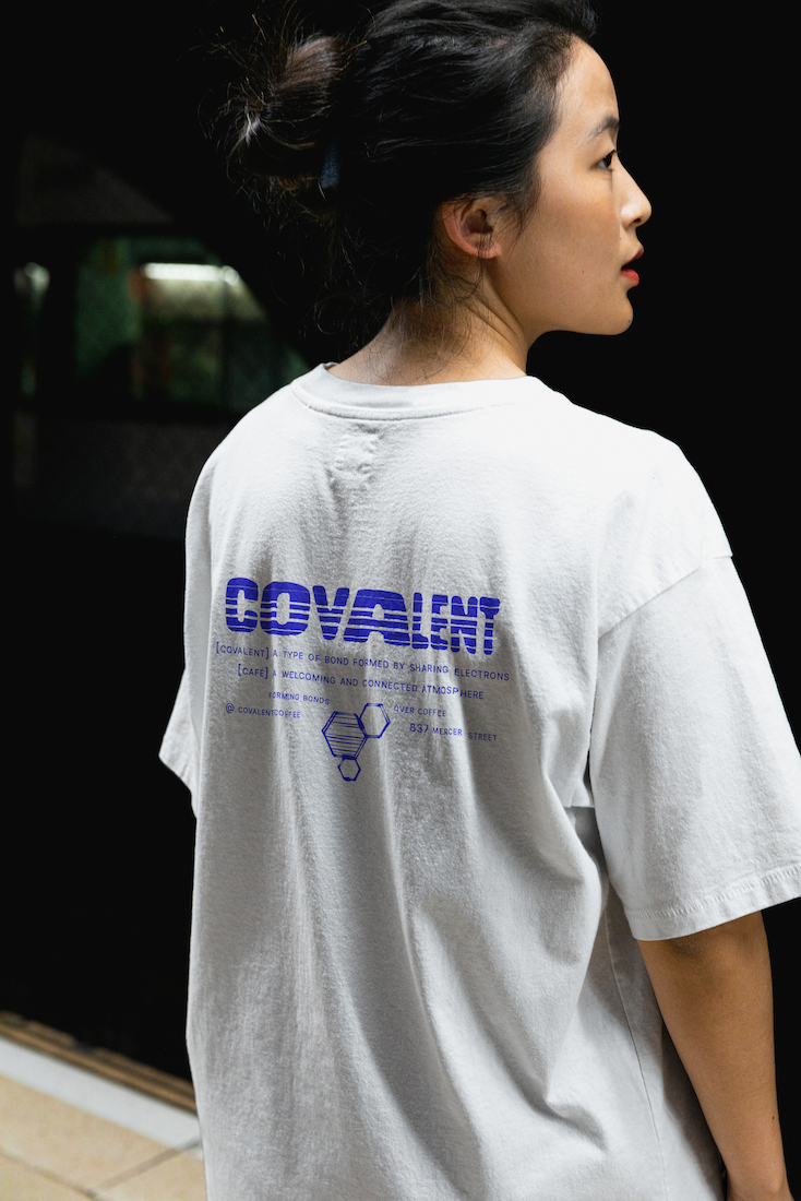A woman with dark hair tied in a messy bun is seen in profile, wearing a white T-shirt with blue text and graphics on the back. The background is dark and out of focus.