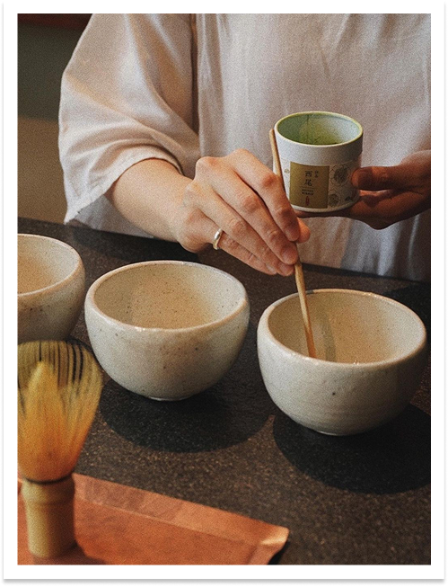 Person preparing to pour tea into ceramic bowls using a bamboo spoon, with other bowls on a dark countertop.