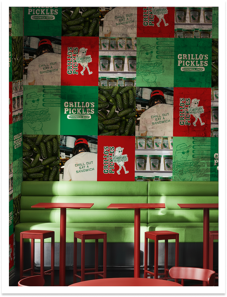 Interior of Grillo's Pickles sandwich shop with a green bench, red tables and stools, and a wall decorated with images of pickles, product packaging, and the shop's mascot, a pickle with sunglasses.
