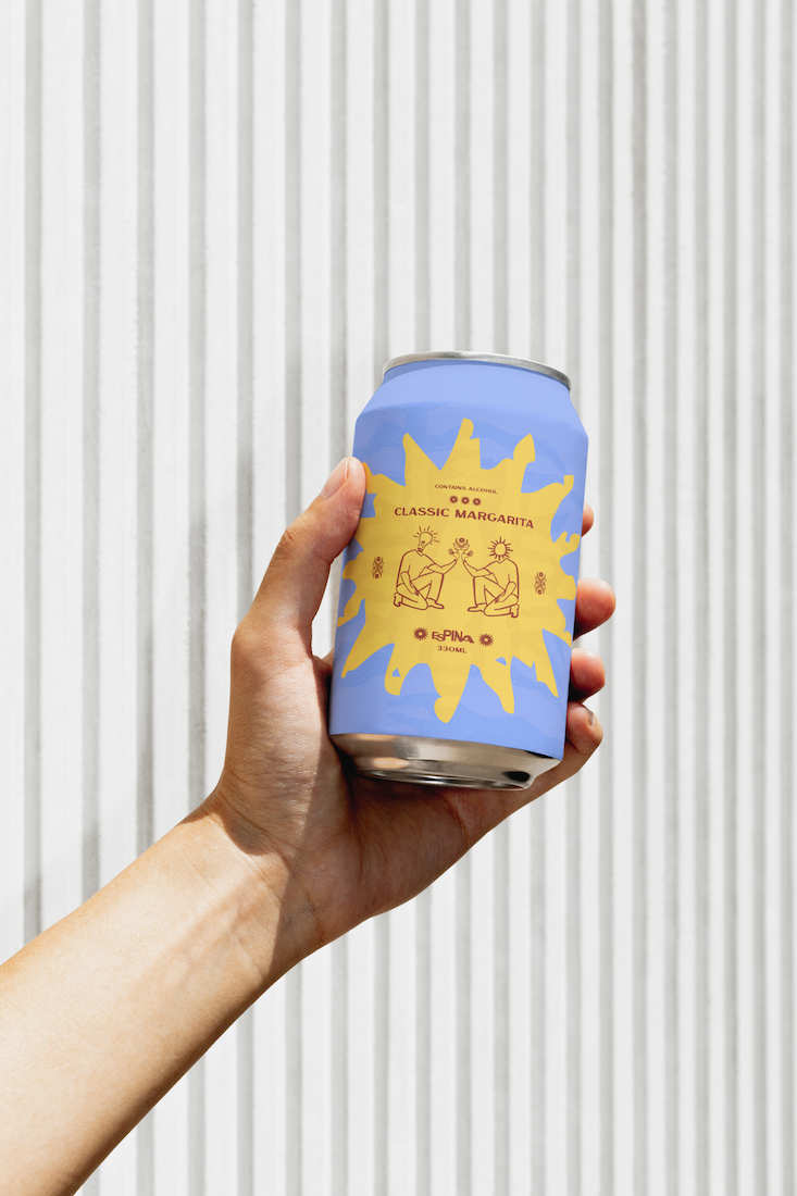 A hand holding a blue can of classic margarita with yellow abstract design and illustrations of two people sitting and drinking together, against a white corrugated metal background.