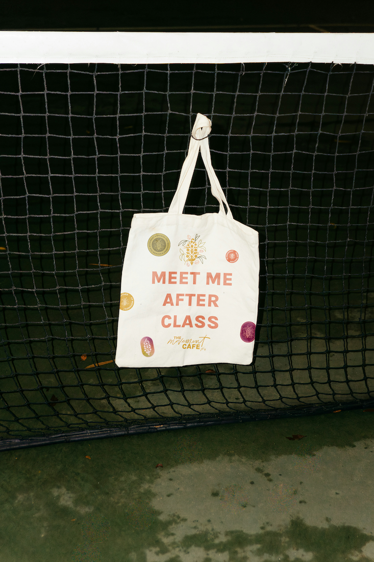 A white tote bag hanging on a black wire fence, with text that reads "Meet me after class" and a small logo below it.