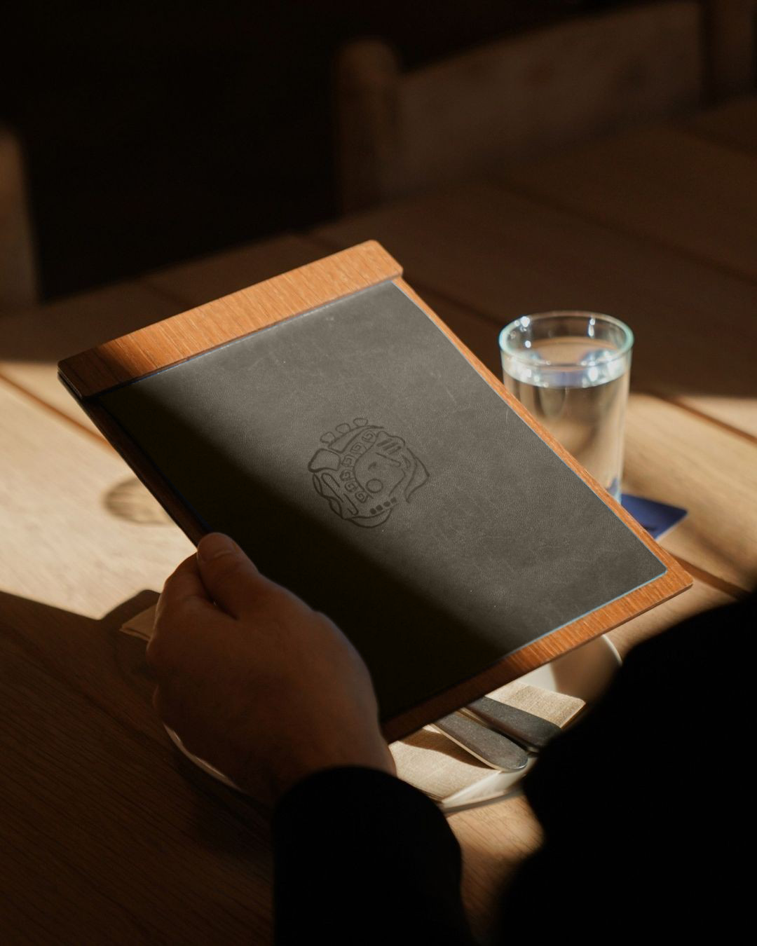 A person holding a menu with a dark cover and a subtle logo, sitting at a wooden table with a glass of water nearby.