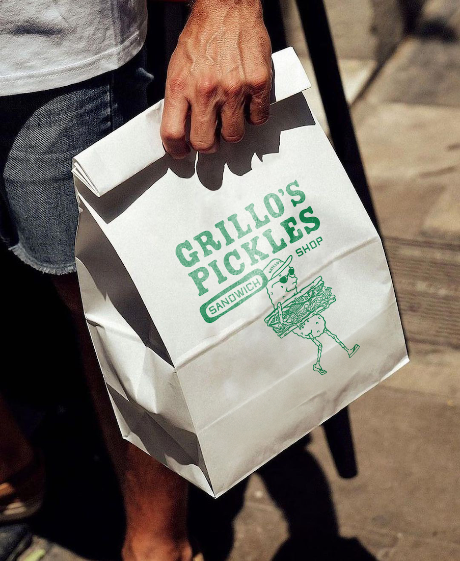 A person holding a white paper bag from Gillo's Pickles Sandwich Shop, featuring a cartoon pickle wearing sunglasses, a hat, and high heels.