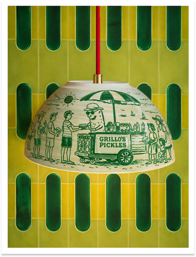 A hanging lampshade with a vintage green and white illustration of a beach scene, featuring a vendor cart labeled "Grillo's Pickles," people in summer clothing, sailboats, and the sun, with a green and yellow tiled background.