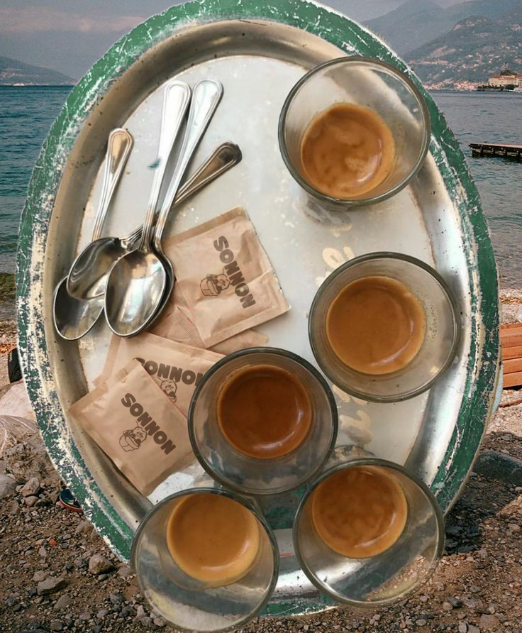 A metal tray with six small glasses of espresso, two sugar packets, and three teaspoons, with a waterfront and mountainous landscape in the background.