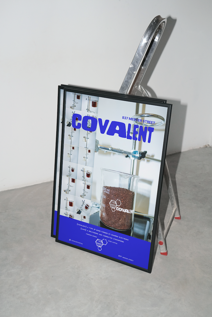 A retractable A-frame signboard displaying promotional material for Covalent, featuring lab equipment and a beaker of ground coffee, placed against a white wall on a concrete floor.