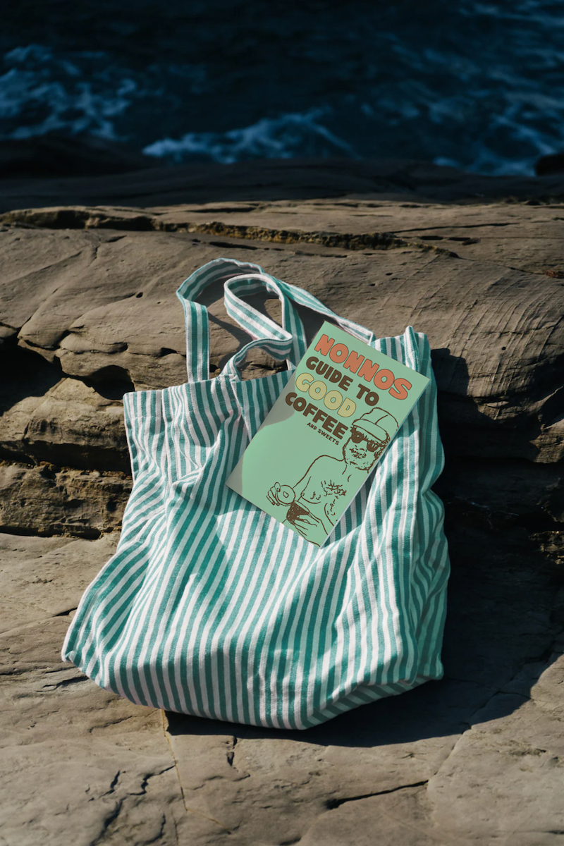 A striped beach bag resting on rocks near the water, with a guidebook titled 'NONNOS Guide to Good Coffee and Sweets' inside.