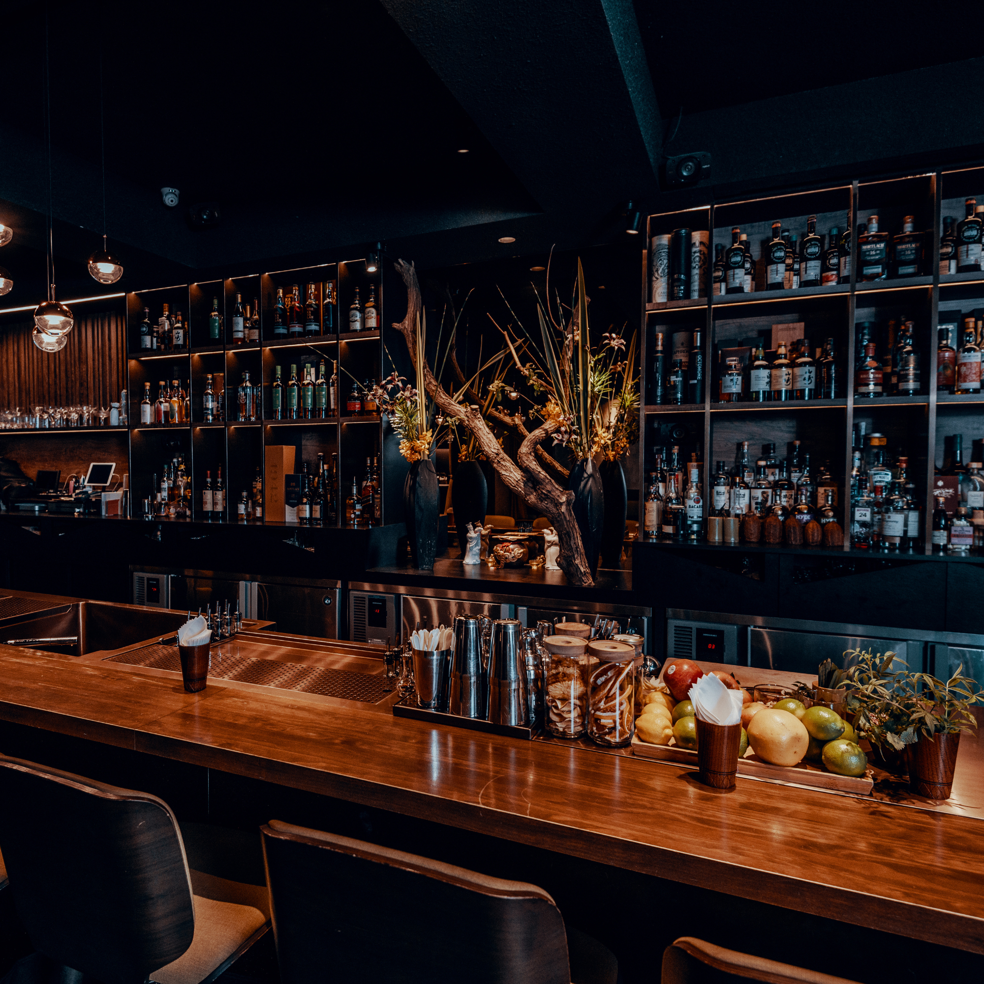 Hotel bar with shelves of liquor bottles, a wooden counter with cocktail glasses, a bowl of apples, and decorative plants, illuminated by warm lighting.