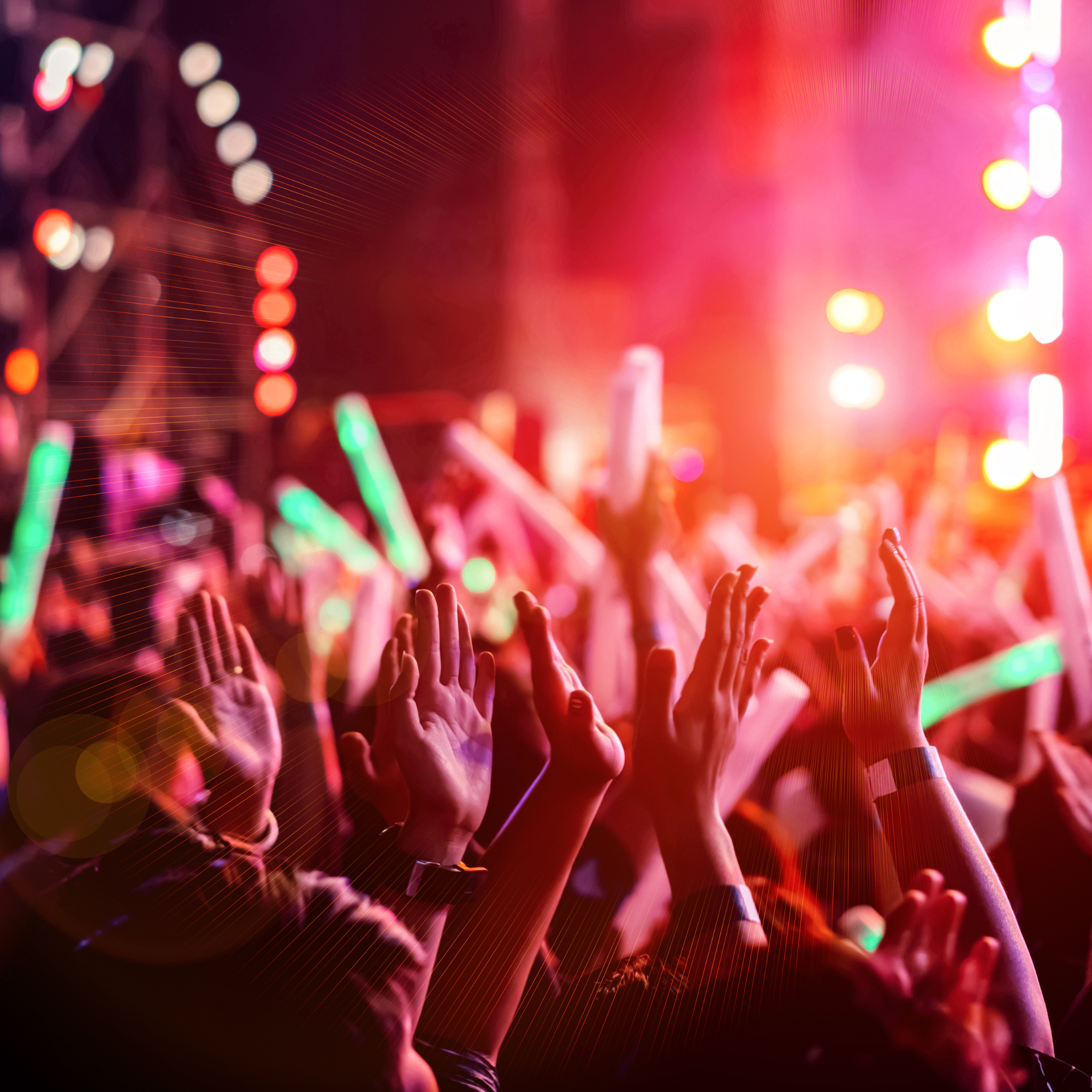 A large crowd of people at a concert or festival with hands raised in the air, colorful lights, and blurred background.