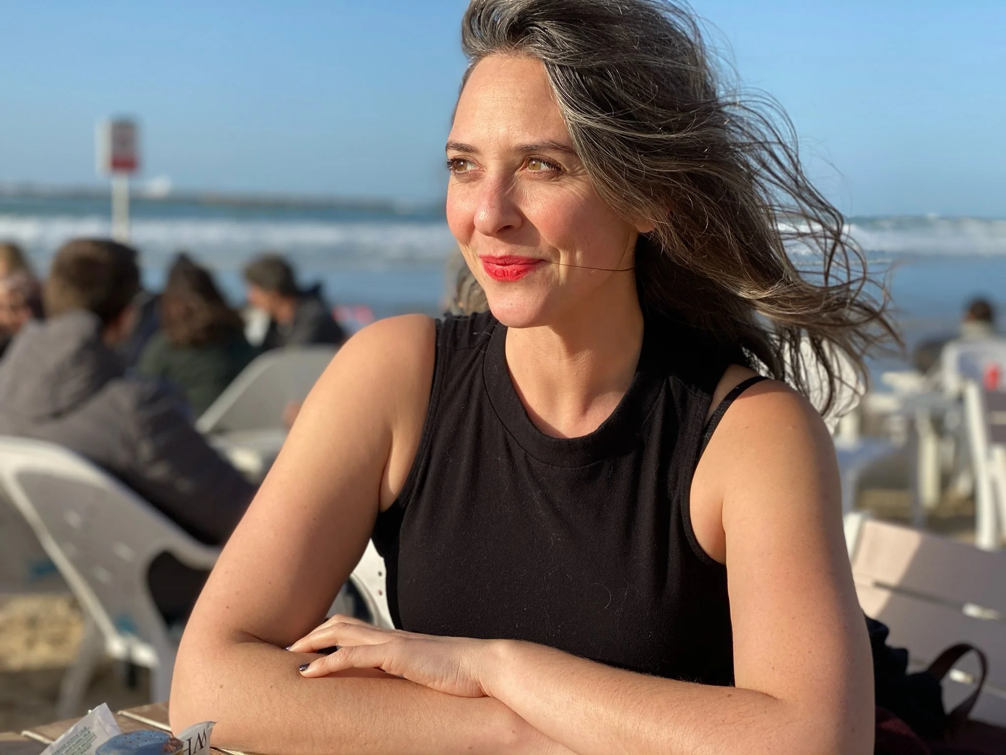 A woman with gray hair and brown eyes sitting at a beachside cafe, smiling and looking to her left, with beach chairs and the ocean in the background.
