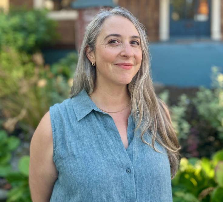 Image of Rachel Toback standing in front of a building with plants. They are a middle aged femme with grey hair.
