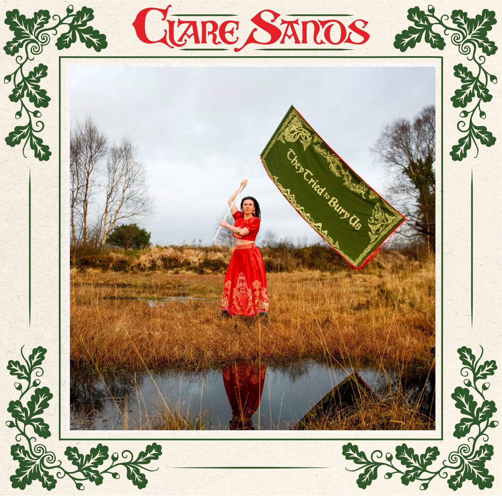 Woman (Clare Sands) standing in a flooded field in a red tribal dress holding a flag above her head