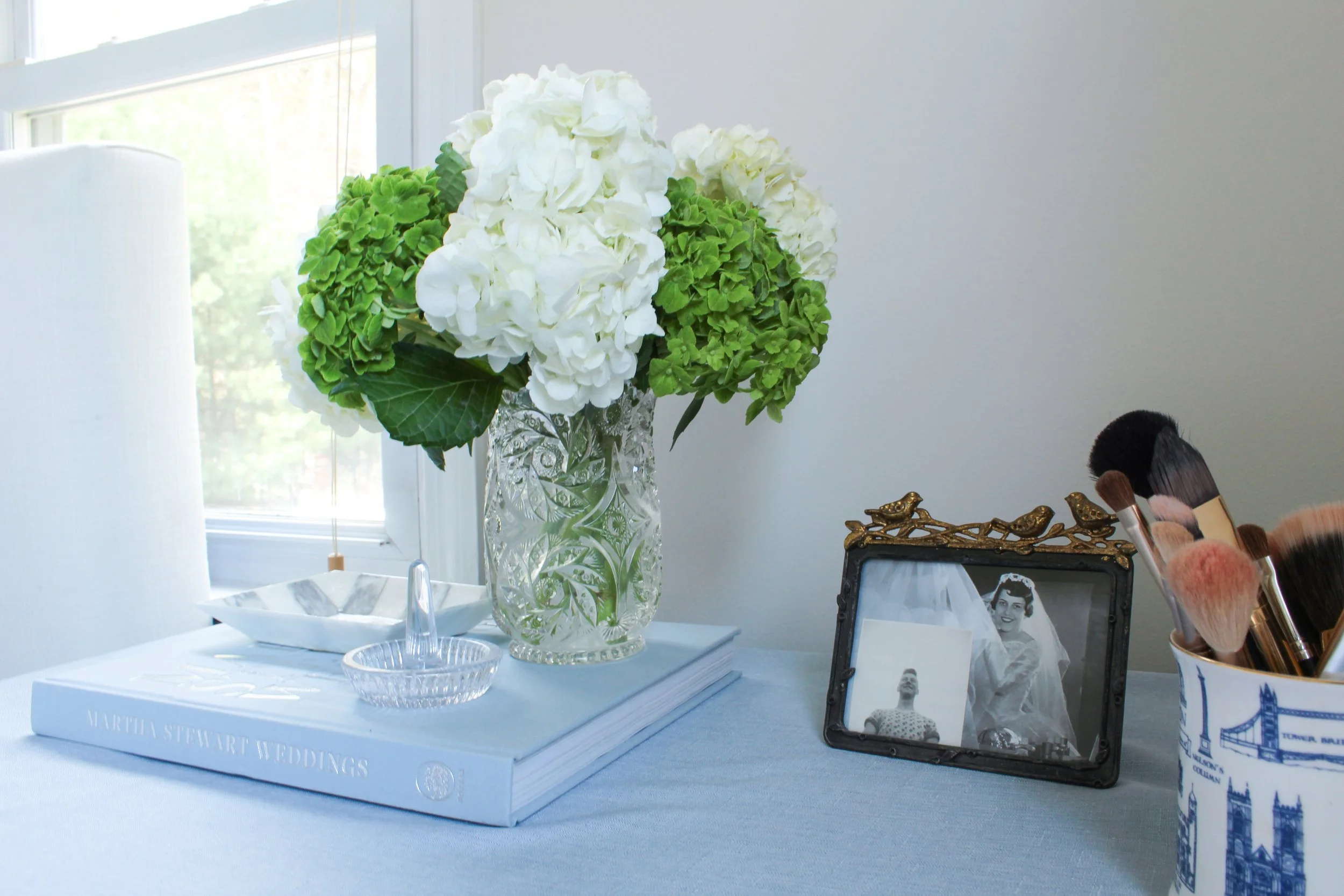 A large bouquet of white and green hydrangeas in a decorative glass vase sits on a white table, with a window in the background and makeup brushes in a patterned container nearby.
