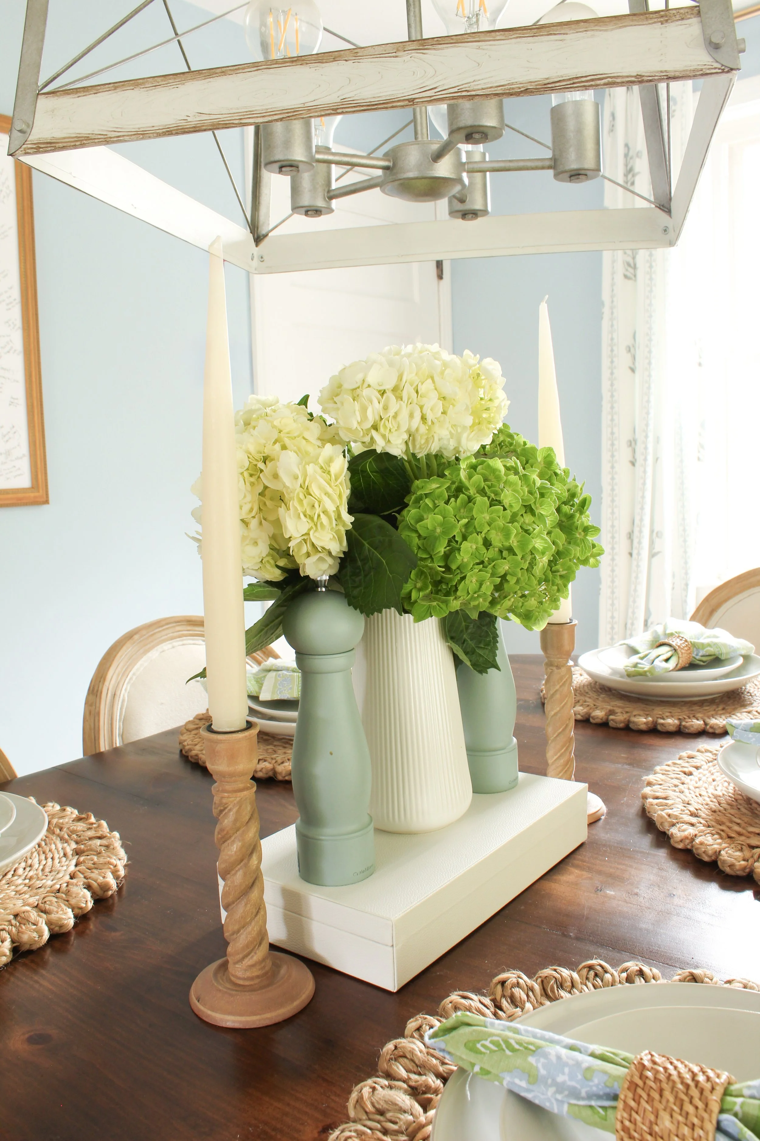 A dining table decorated with a white wooden and metal chandelier, a central flower arrangement with white hydrangeas and green flowers in pastel-colored vases, surrounded by woven placemats, plates, napkins, and chairs with woven backs in a bright room with natural light.
