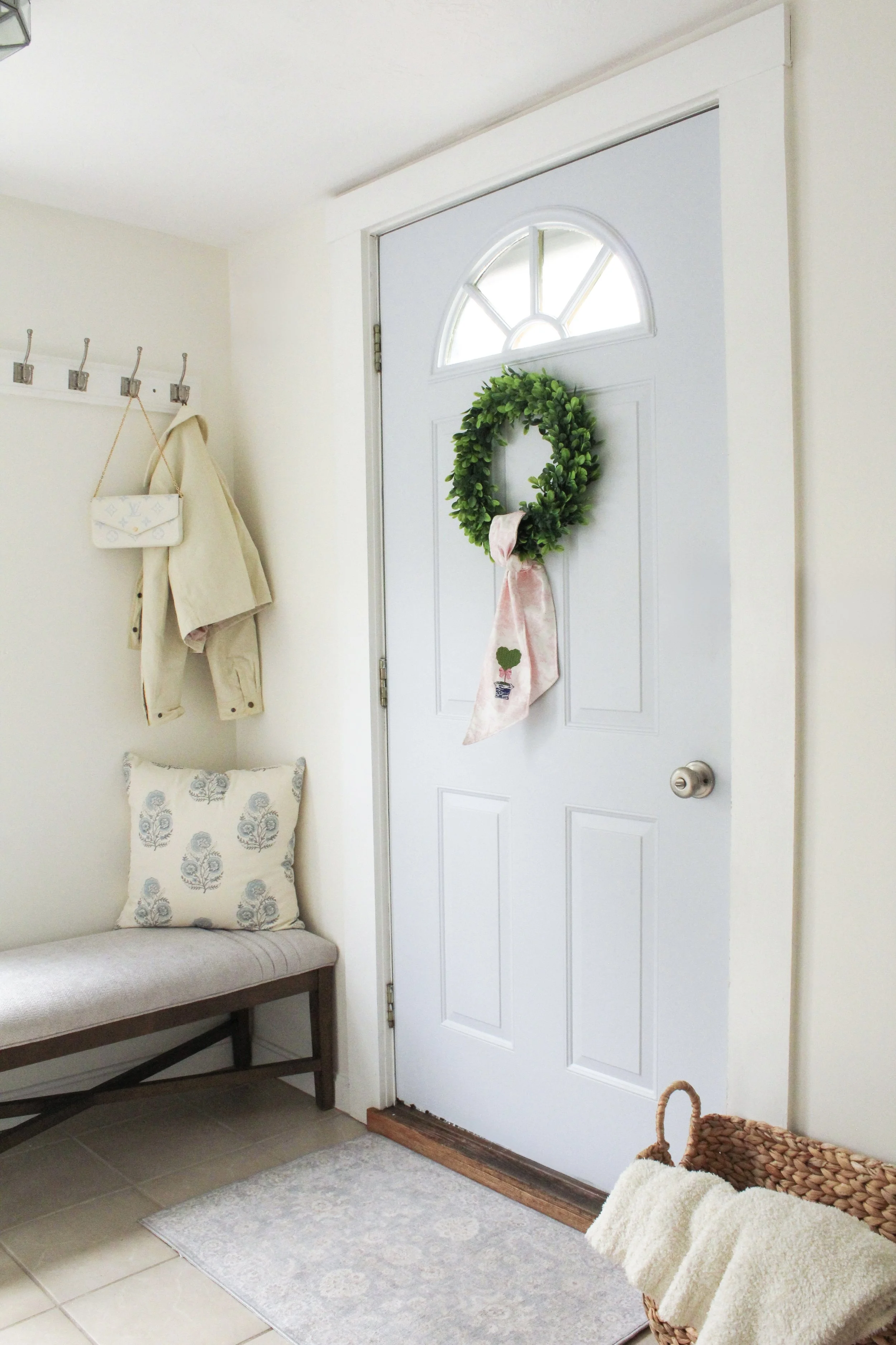 Entryway with a white door decorated with a green wreath and a pink ribbon. A coat rack with hooks on the wall with jackets and a small white purse hanging. A cushioned bench beneath the coat rack with a decorative pillow. A woven basket with a towel or blanket on it. A light-colored rug on tile flooring.