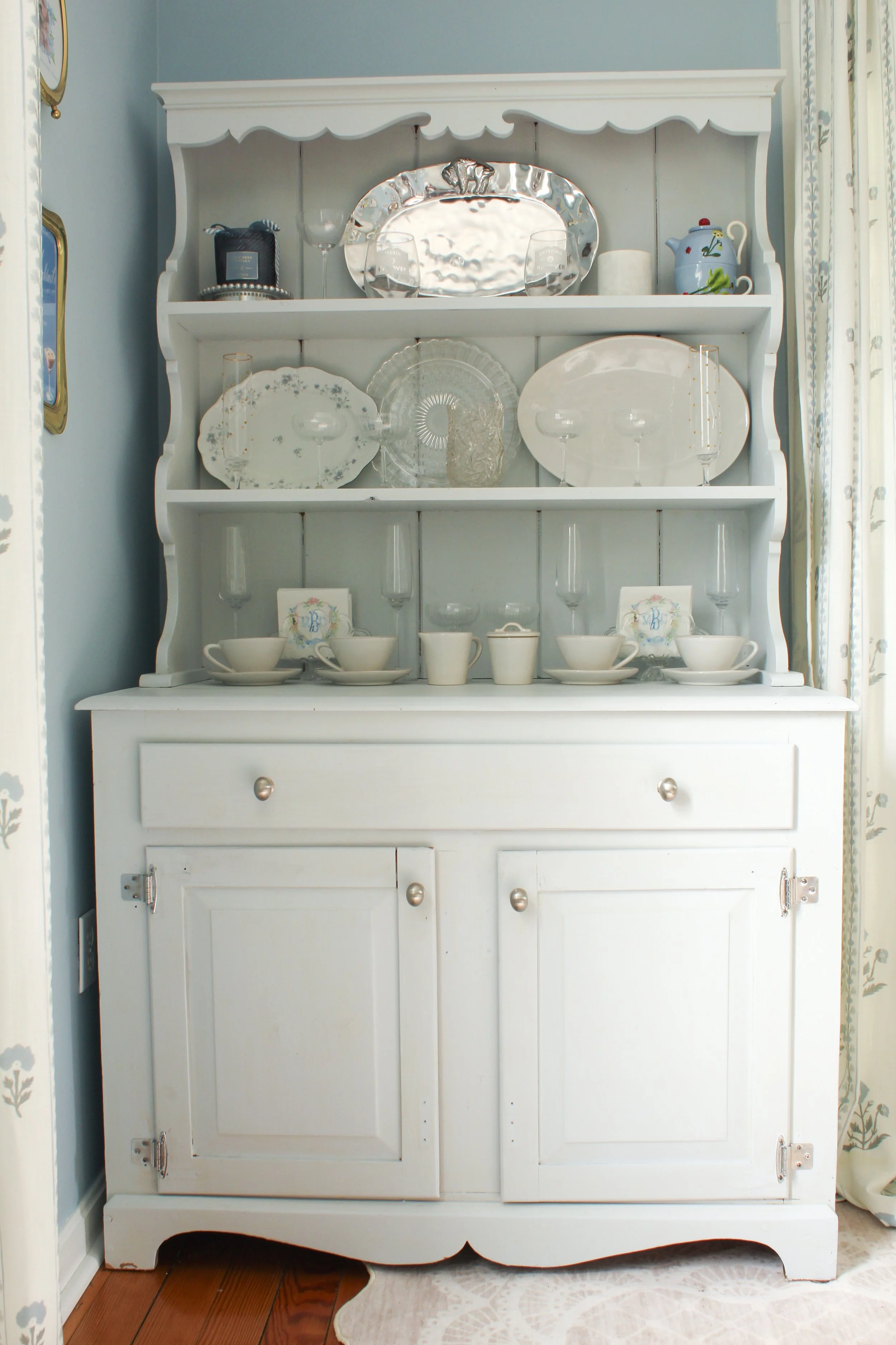 White wooden hutch displaying glassware, plates, cups, and teapot, situated near a window with floral curtains.