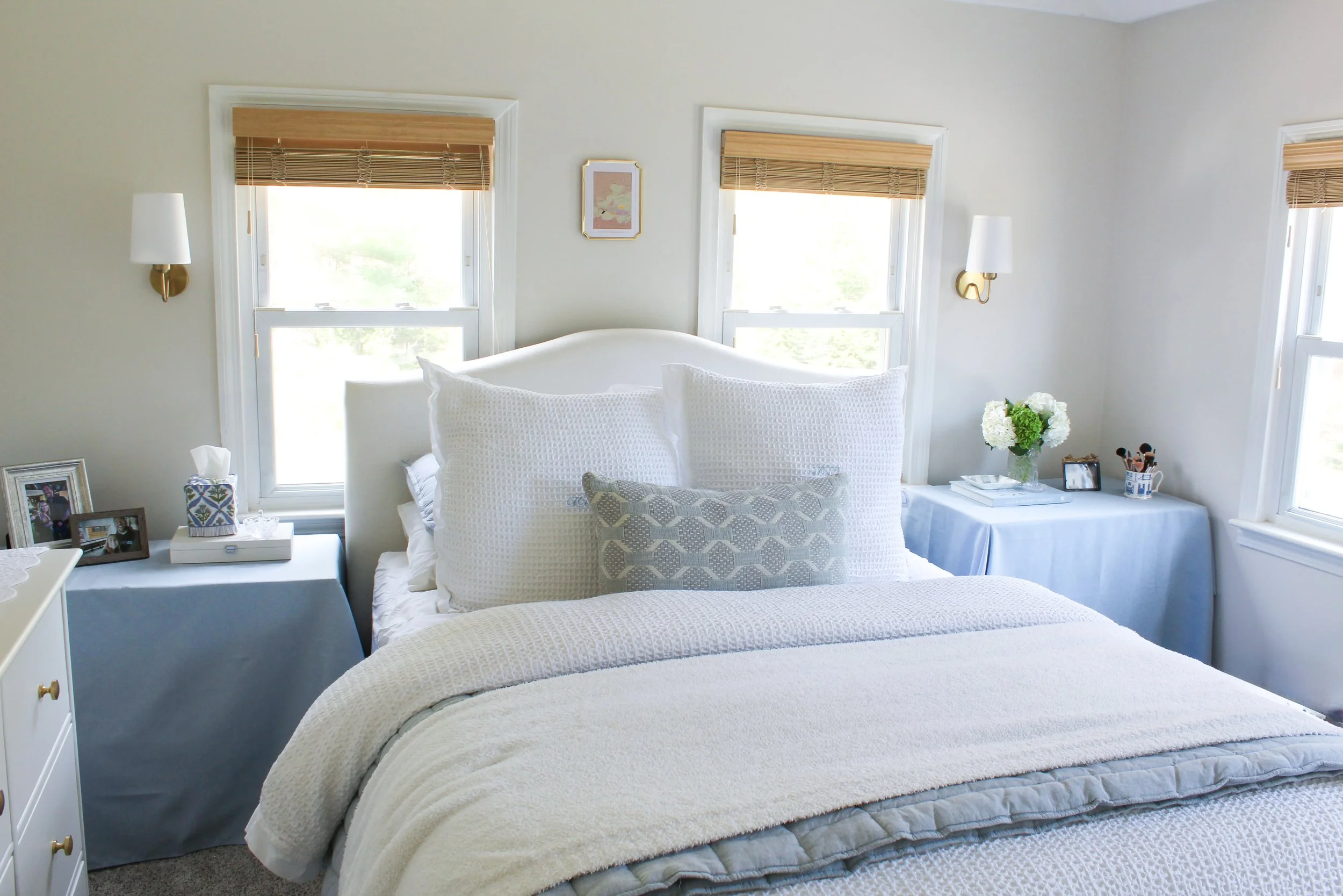 A cozy bedroom with white walls, three windows with bamboo blinds, a white bed with textured pillows, two wall-mounted lamps, a nightstand with flowers, framed photos, and a small tray.