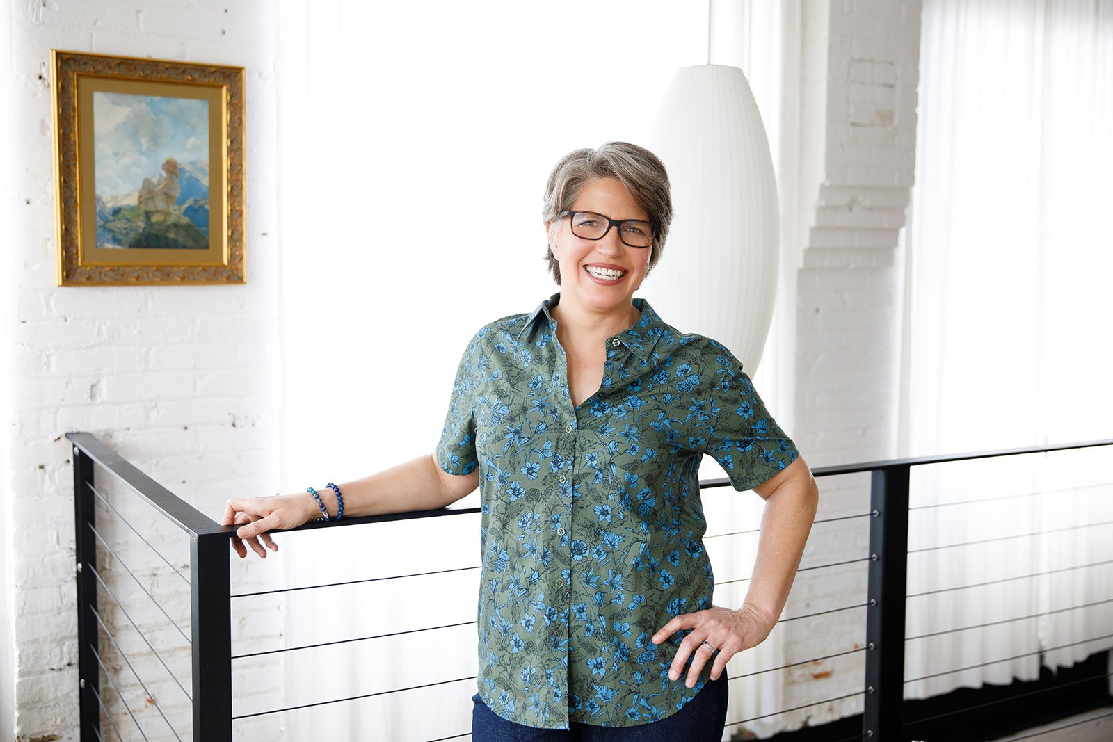 A smiling woman with glasses and short gray hair stands indoors, leaning on a black railing with her left hand resting on it and her right hand on her hip, wearing a green shirt with blue floral pattern. The background features a white brick wall, a painting in a gold frame, and a large white lamp with natural light streaming in through a window.