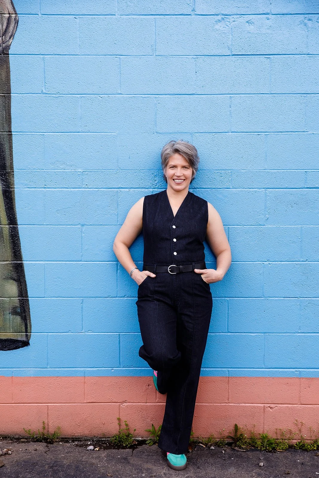 Woman with short gray hair, wearing a sleeveless black vest and black pants, standing against a colorful blue and pink painted brick wall, smiling at the camera.