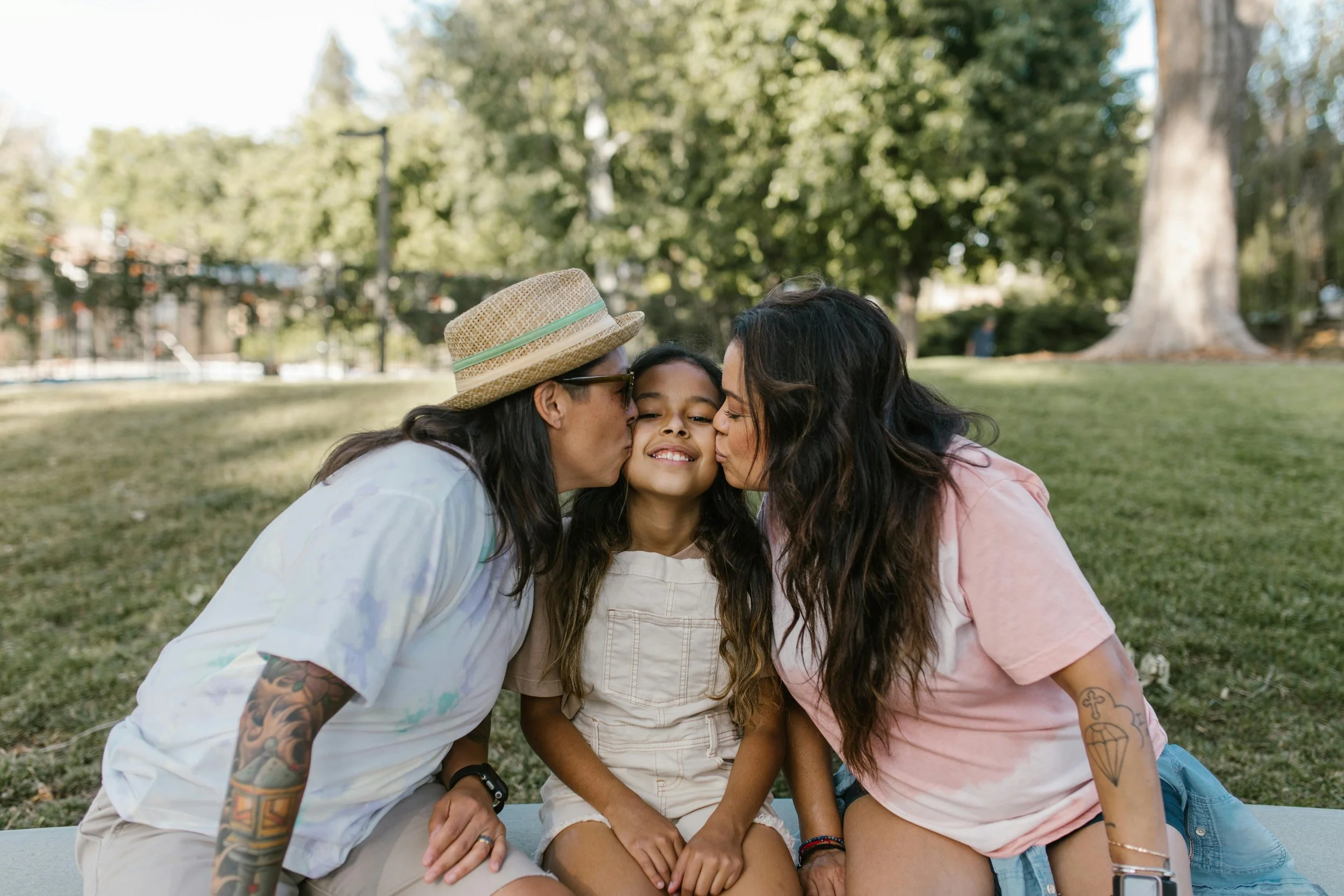 Three people, two women and a girl, sharing a kiss on each cheek in a park with green trees in the background.