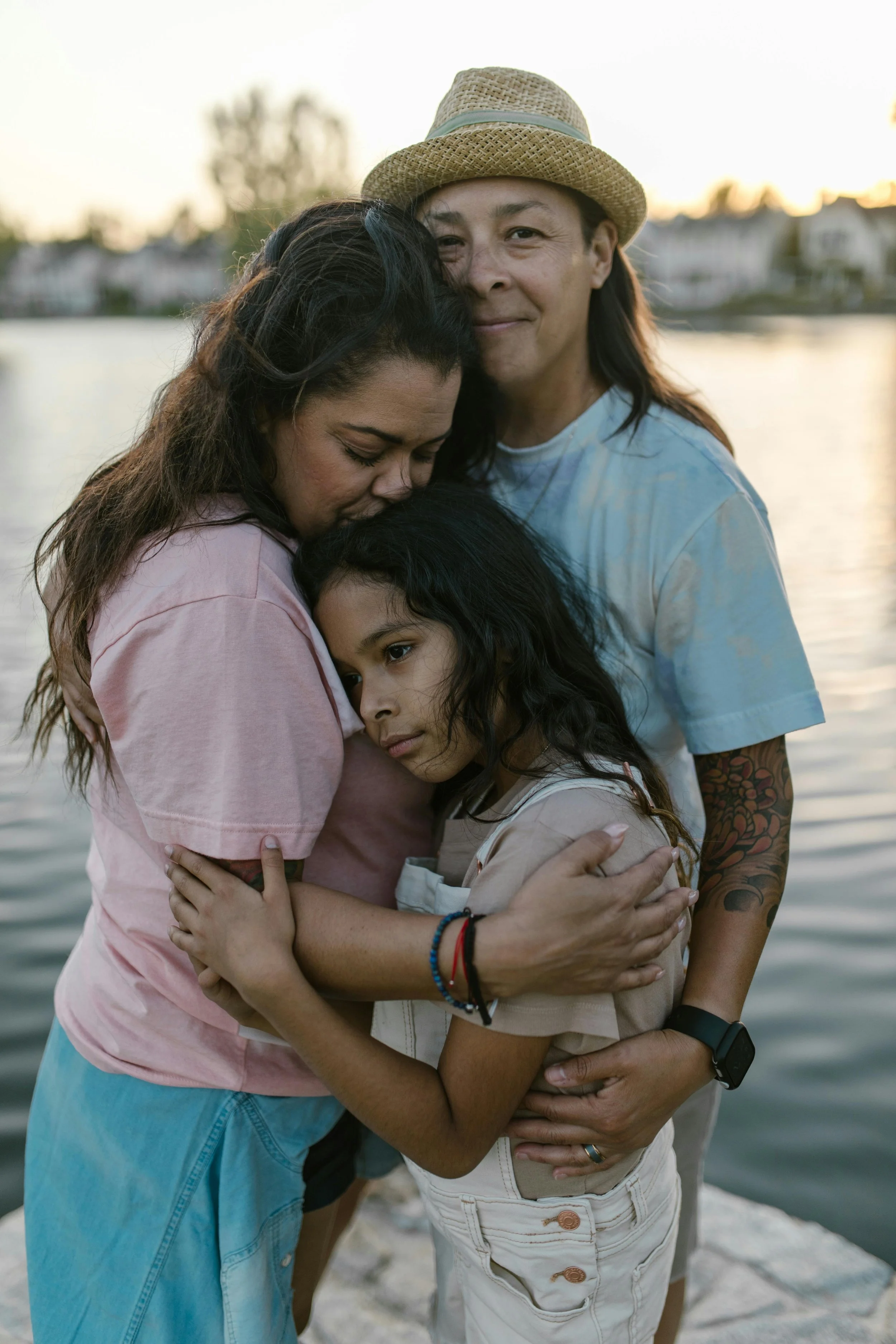 Family hugging by a body of water during sunset, with houses and trees in the background.