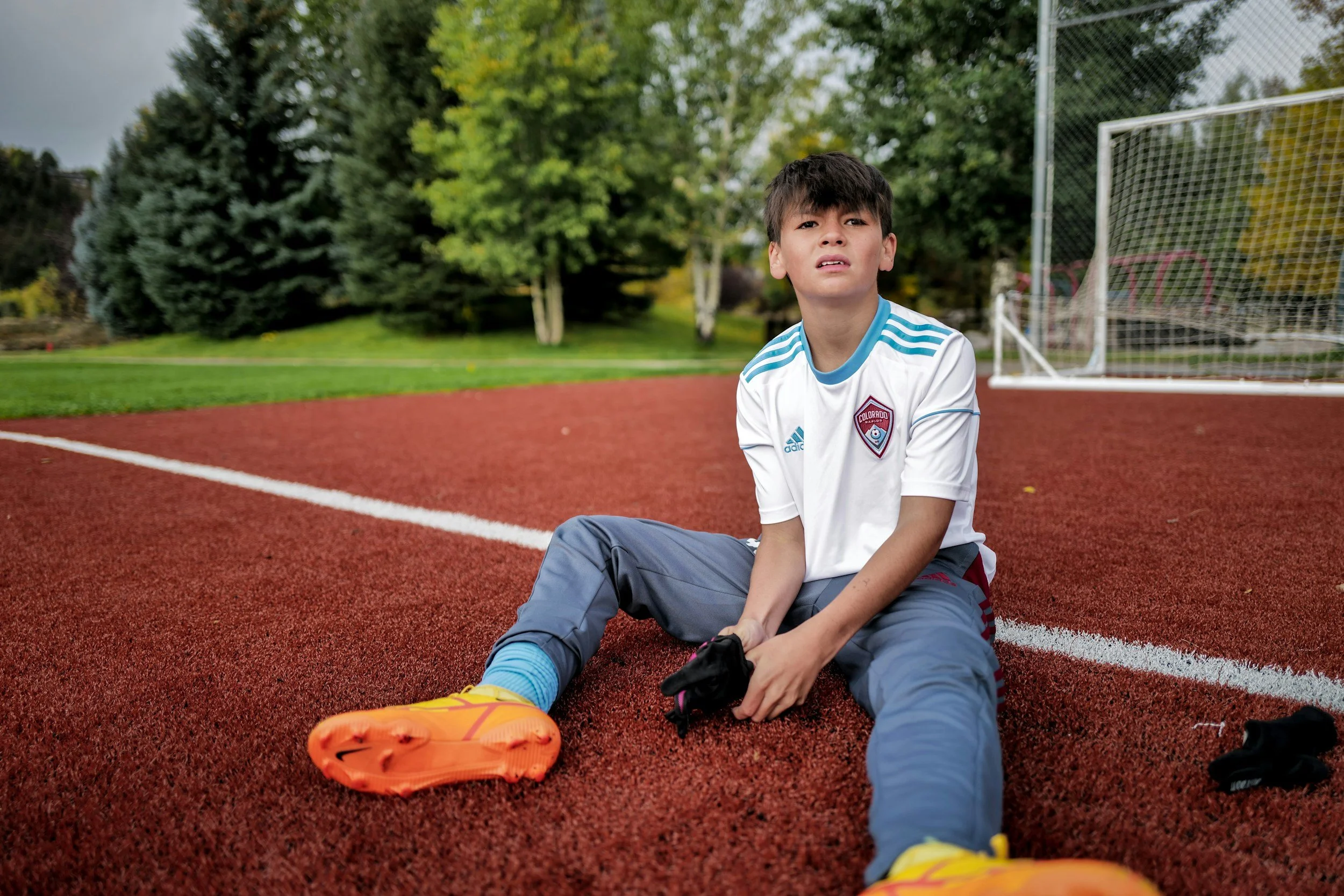 Young boy sitting on a red running track, stretching or tying his shoes, with trees and a soccer goal in the background.
