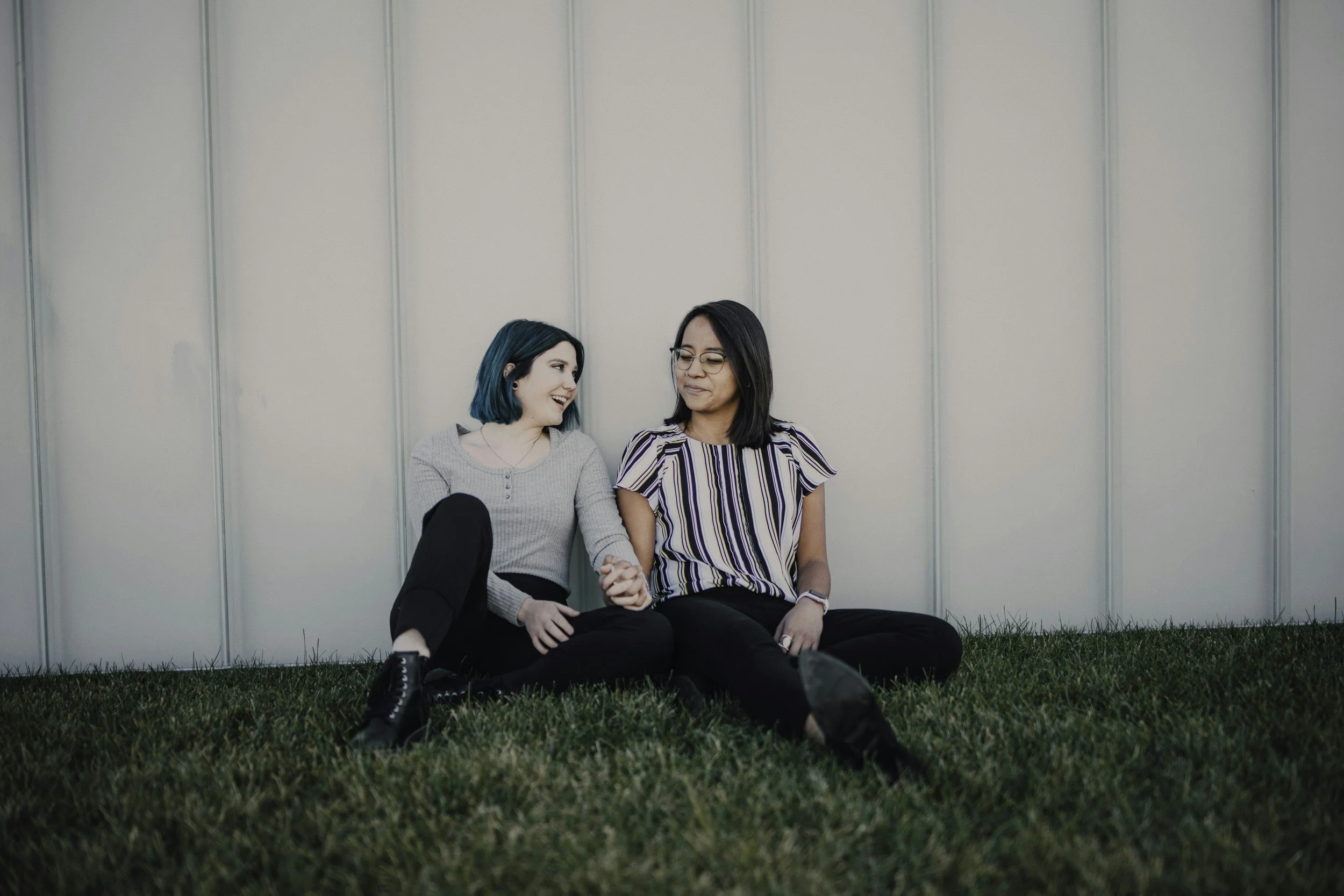 Two women sitting on grass against a metal wall, holding hands and smiling at each other.
