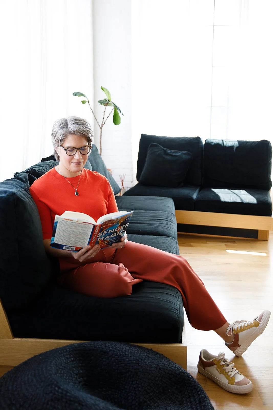 A woman sitting on a black couch reading a book in a bright living room, wearing glasses, a red top, orange pants, and white sneakers with yellow accents, with a potted plant and a wooden floor in the background.
