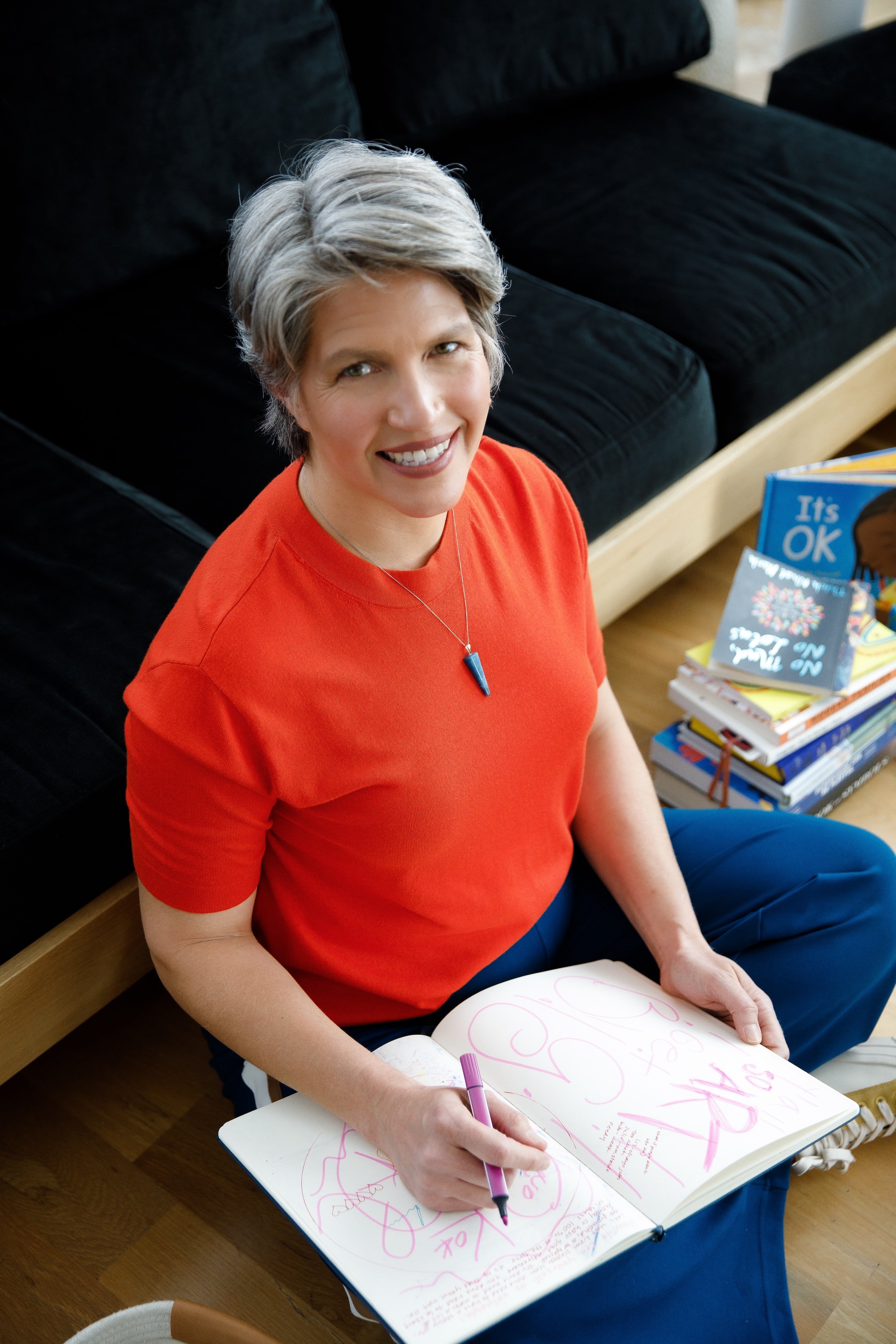 A woman with short gray hair, wearing an orange shirt and blue pants, sitting on the floor, writing in a large sketchbook with pink and purple markers. She is smiling and looking up at the camera. In the background, there are books and a black couch.