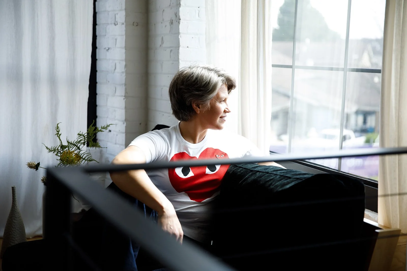 A woman with short gray hair looking out a large window, smiling. She is wearing a white t-shirt with a red heart and black dots. She is sitting on a black sofa in a bright room with white curtains, a white brick wall, and a plant in the background.