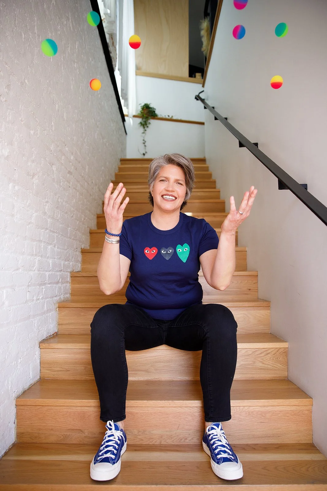 A woman with short gray hair sitting on wooden stairs, smiling and gesturing with her hands. She is wearing a navy blue t-shirt with colorful heart designs, black jeans, and blue sneakers. The staircase has white brick walls on the sides, a black handrail on the right, and some hanging colorful balloons in the background.