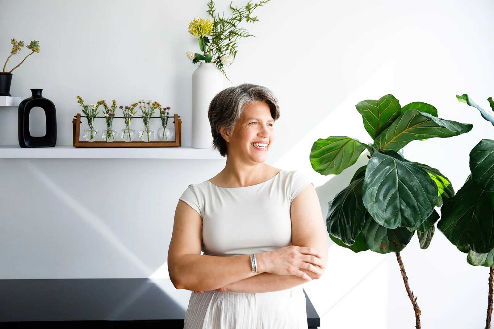 A woman with short gray hair wearing a white top smiling and looking to her right in a bright, minimalistic room with large green plant, white shelves, and decorative vases.