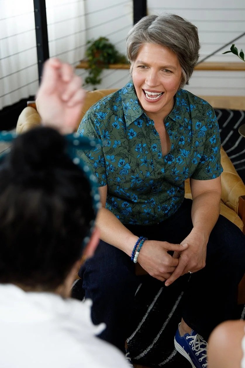 A woman with short gray hair laughing and talking to someone. She is wearing a green shirt with blue floral patterns, seated on a tan couch, with a plant in the background.