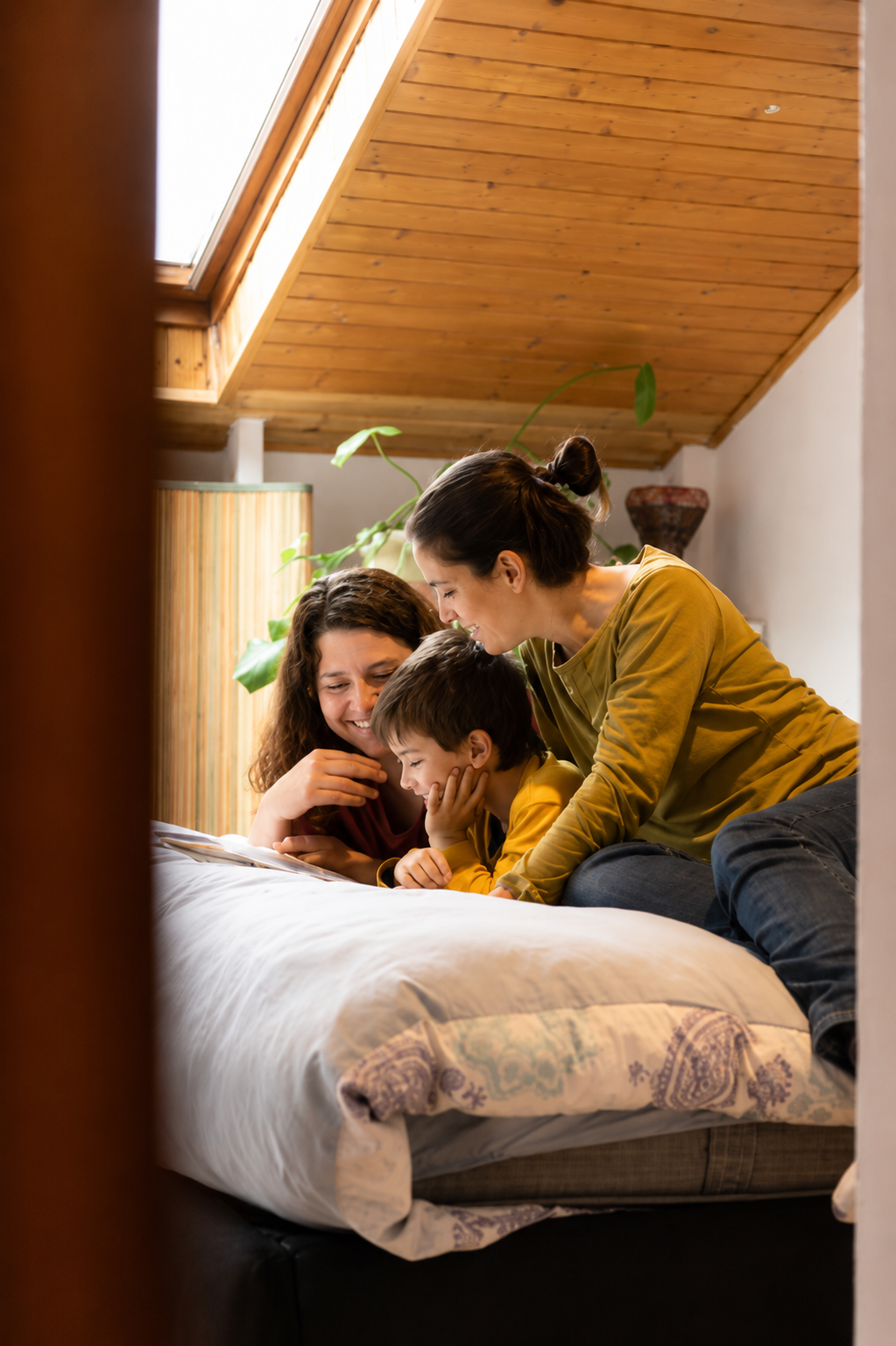 Three people, two women and a young boy, sitting on a bed in a cozy room with a wooden ceiling. They are smiling and looking at something together, enjoying a happy moment.