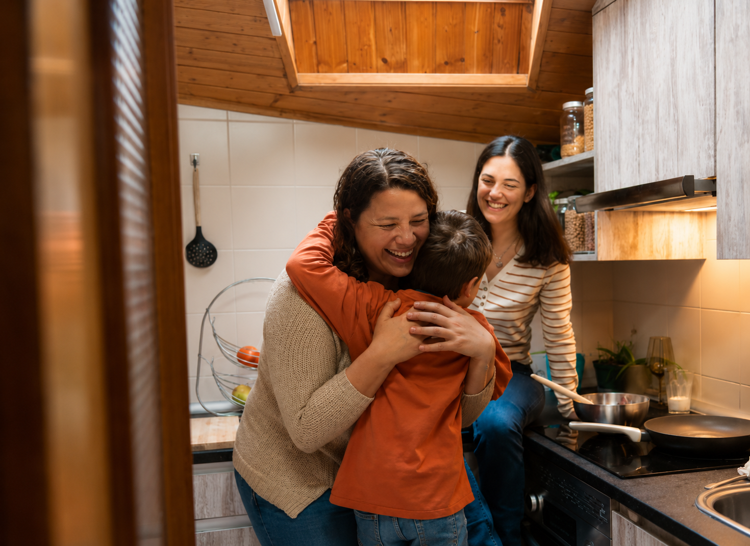Three people, two women and a boy, hugging and smiling happily in a cozy kitchen with wood-paneled ceiling.