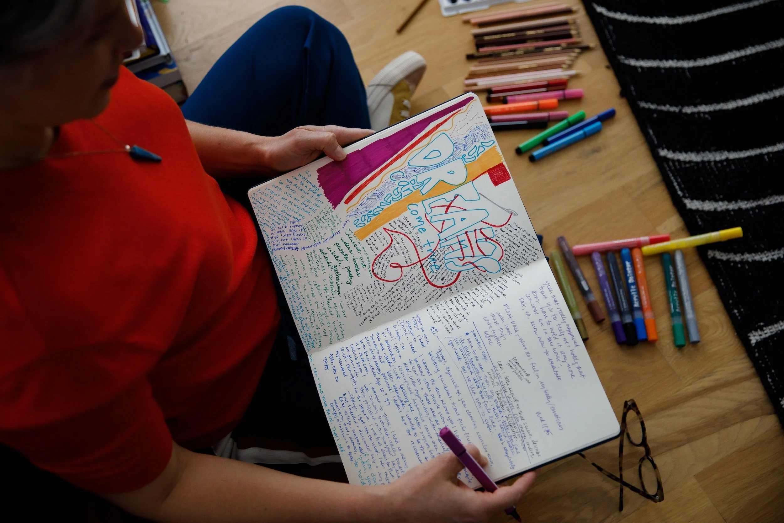 A person in an orange shirt sitting on the floor, reading a colorful, handwritten journal page with blue, red, purple, orange, and green ink. There are various colored markers laid out on the wooden floor next to them, and a black and white striped textile nearby.