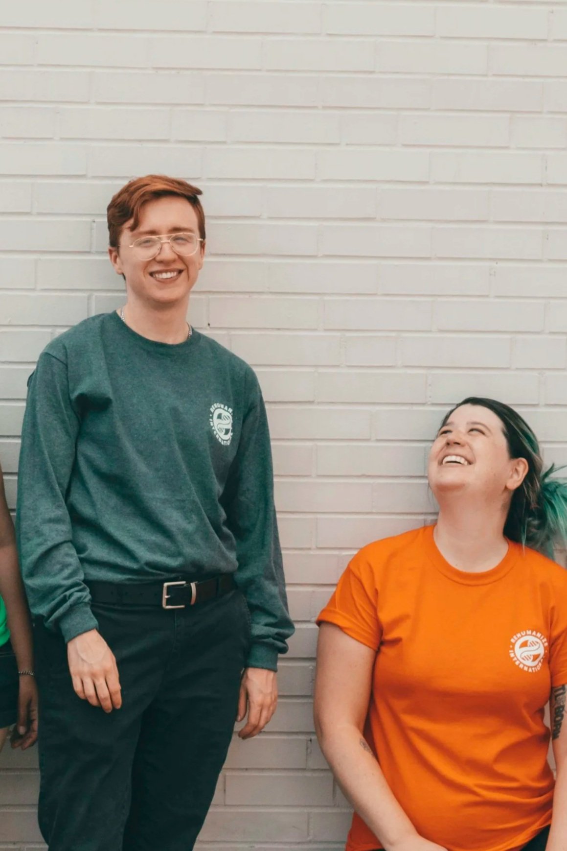 Two people, a man and a woman, are standing against a white brick wall. The man has short reddish-brown hair, glasses, and is smiling, wearing a dark green long-sleeve shirt and black pants. The woman has dark hair with colored highlights and is sitting, looking up and smiling, wearing an orange T-shirt with a logo on the chest.