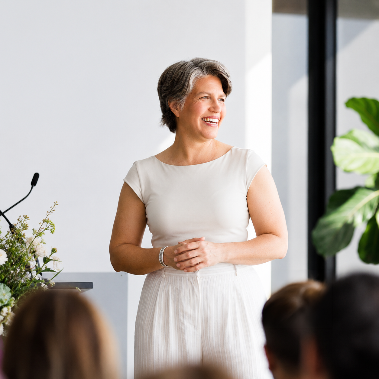 A woman with short gray hair, wearing a white top and light-colored pants, smiling and standing in front of a group, possibly giving a presentation or speech in a bright, modern room with a microphone and plants.