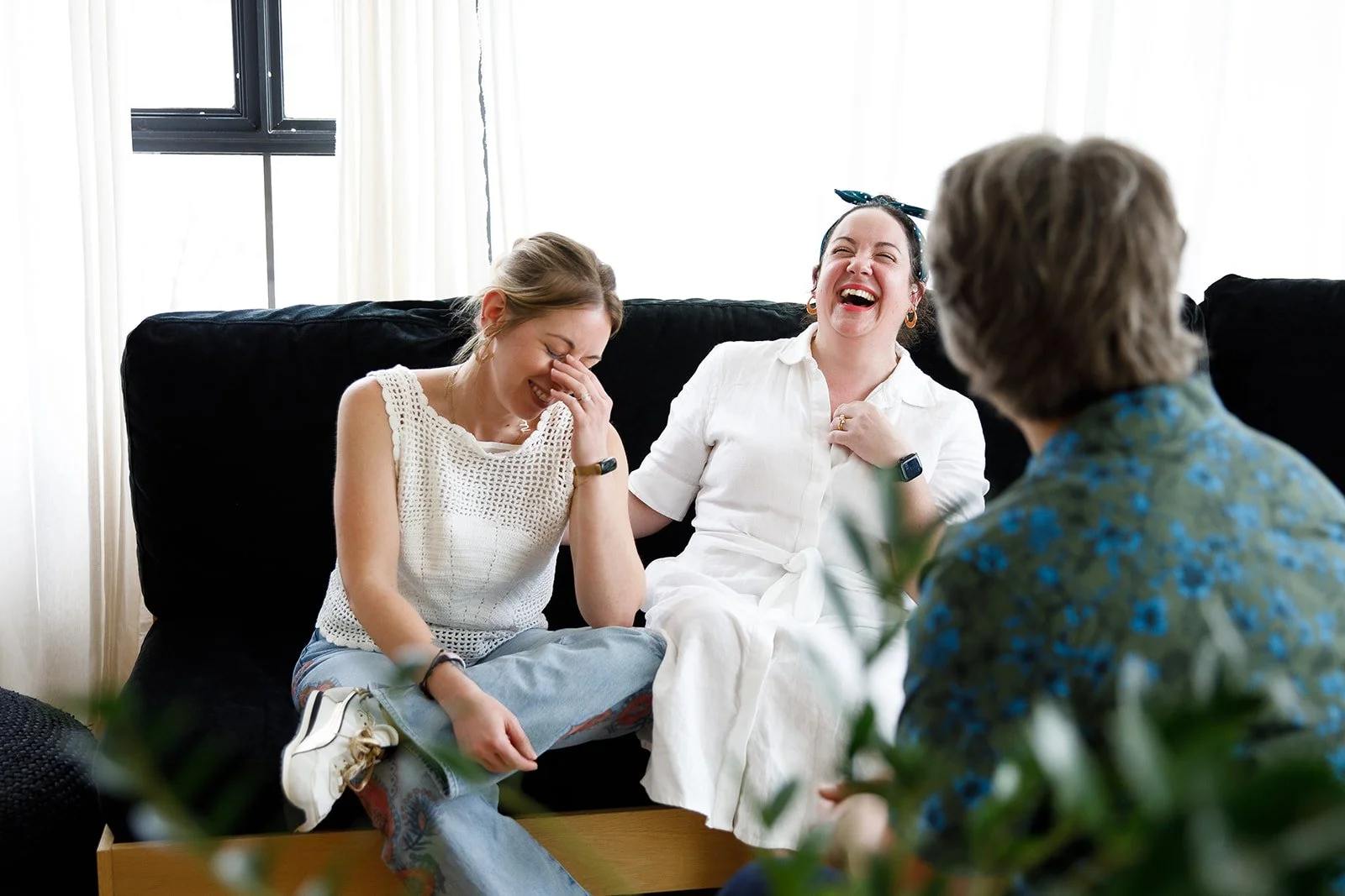 Three women sitting on a black couch, laughing and talking, with a man facing them, in a bright room with white walls and a window.