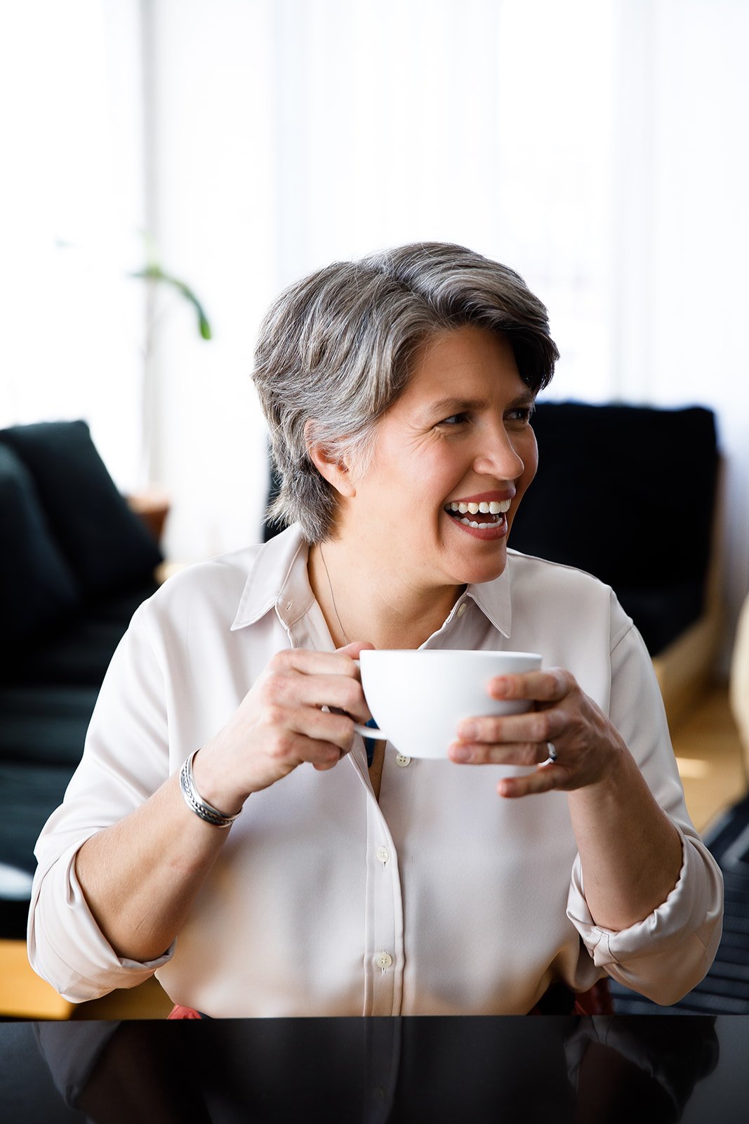 A middle-aged woman with short gray hair smiling and laughing while holding a white mug in a cozy indoor setting.
