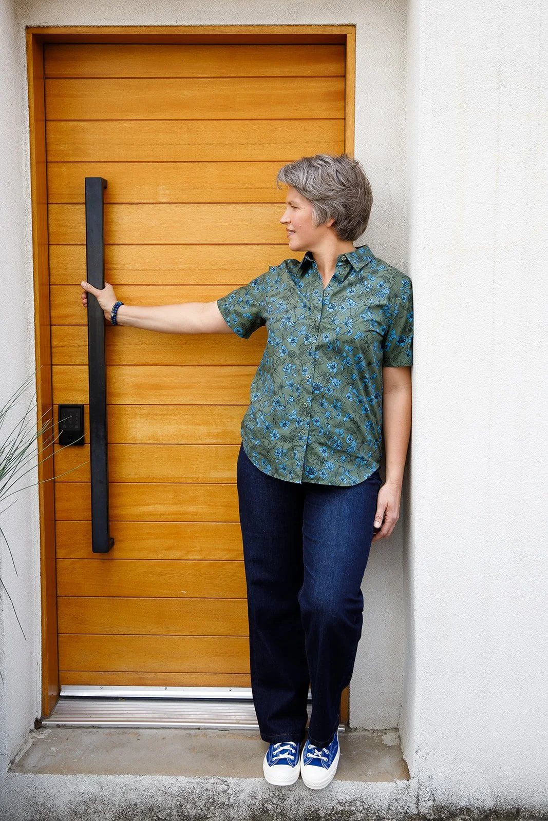 A woman with short gray hair wearing a green floral shirt and dark jeans stands outside a wooden door, holding a black handle and smiling.