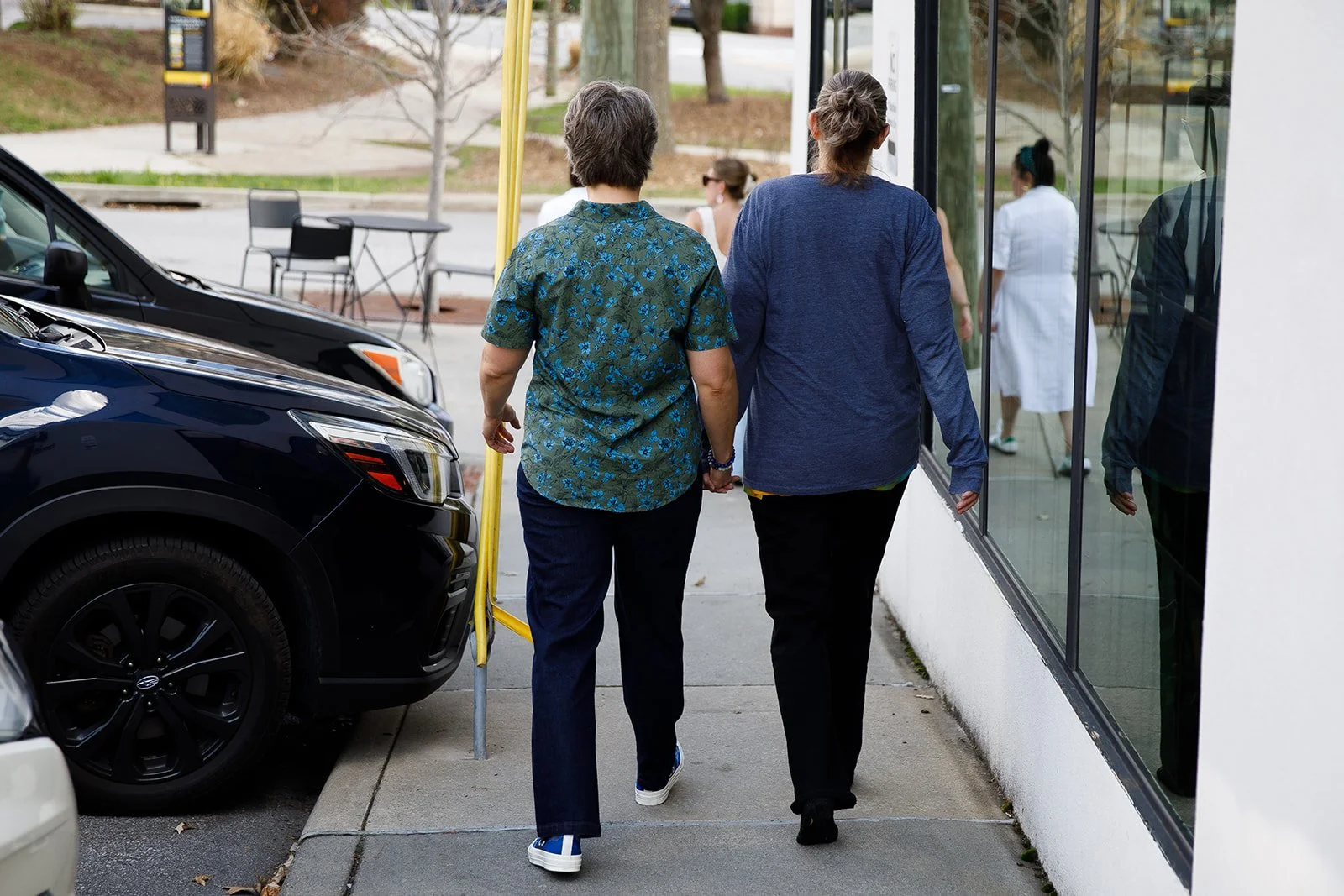 Two women holding hands while walking on a sidewalk outside a building, with cars parked beside them and people visible inside through a glass window.