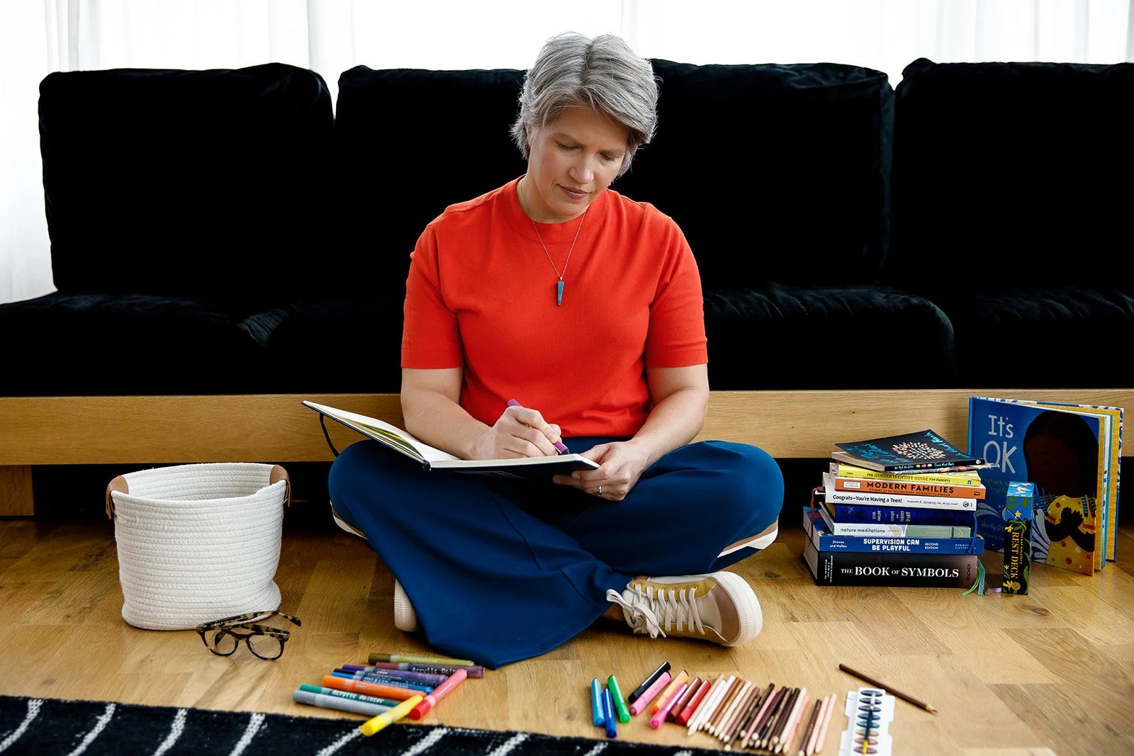 Woman sitting cross-legged on a hardwood floor, writing in a notebook, surrounded by colorful markers, books, and children's items, with a black couch behind her.