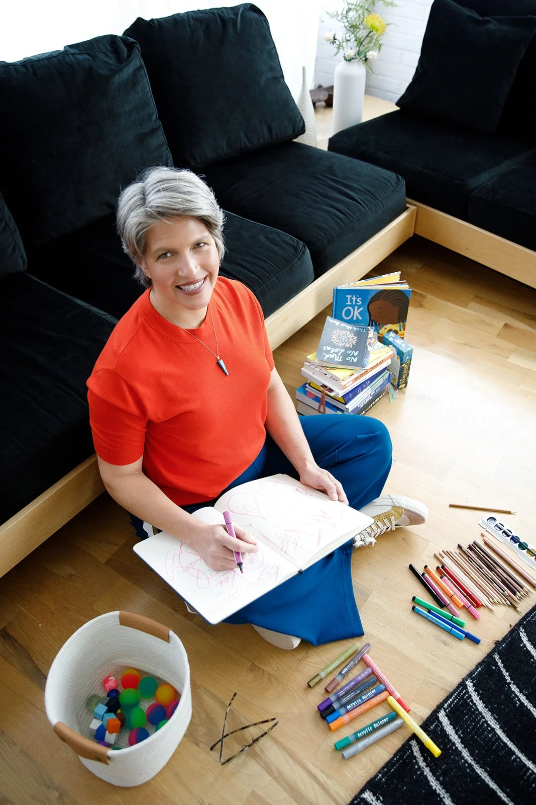 Woman with short gray hair sitting on the floor, drawing in a sketchbook with colorful markers around her, and a stack of children's books nearby.