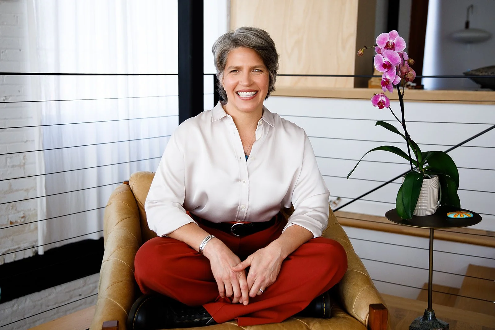 A woman with short gray hair sitting on a tan armchair in a modern indoor setting, smiling, with a pink orchid in a pot on a small black table beside her.